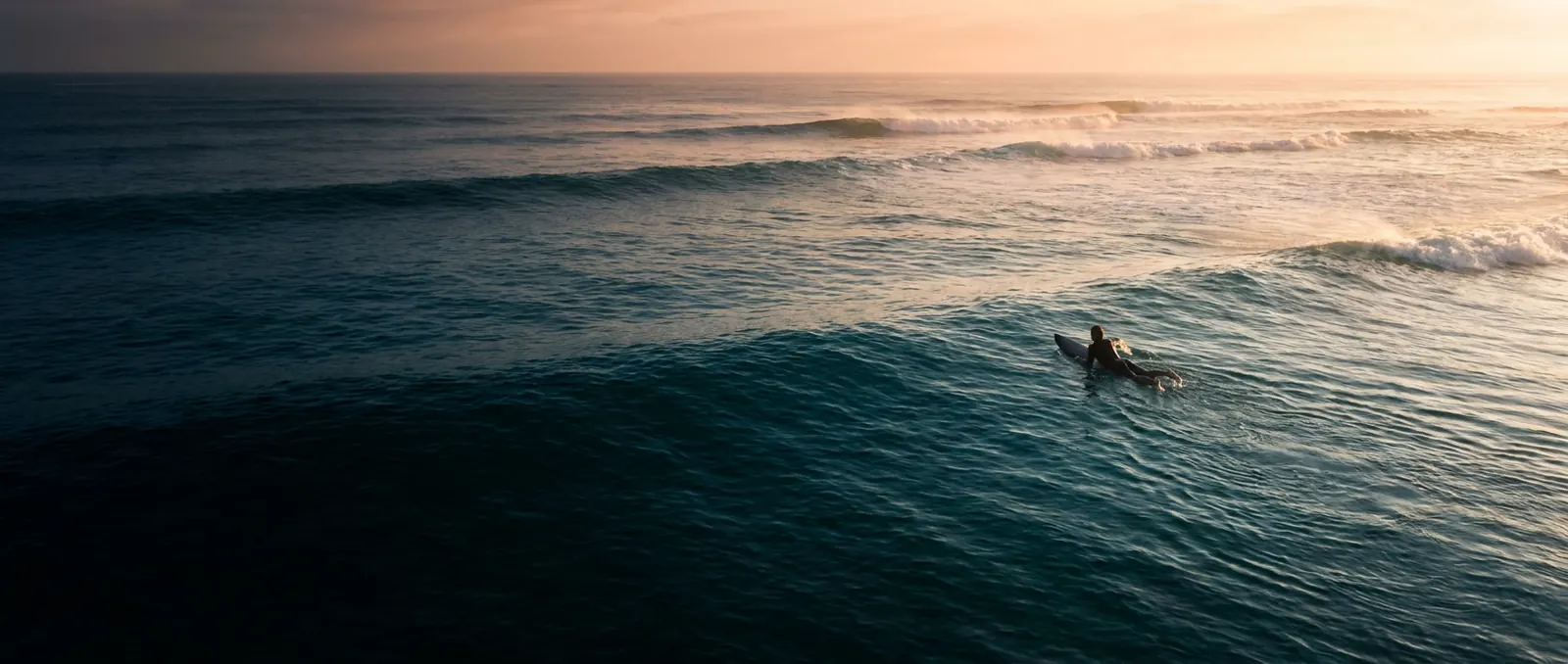 Surfboard volume calculator — a lone surfer paddling a shortboard out through glassy dawn swell